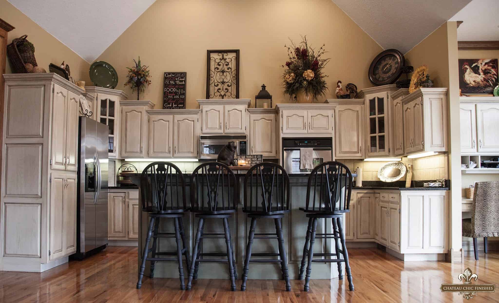 Spacious kitchen with high ceilings, cream-colored cabinets enhanced by white paint for a fresh look, and wooden floors. Center island with four black wooden bar stools. Decorative plants and framed art above cabinets. Stainless steel refrigerator and built-in oven. Warm lighting throughout.