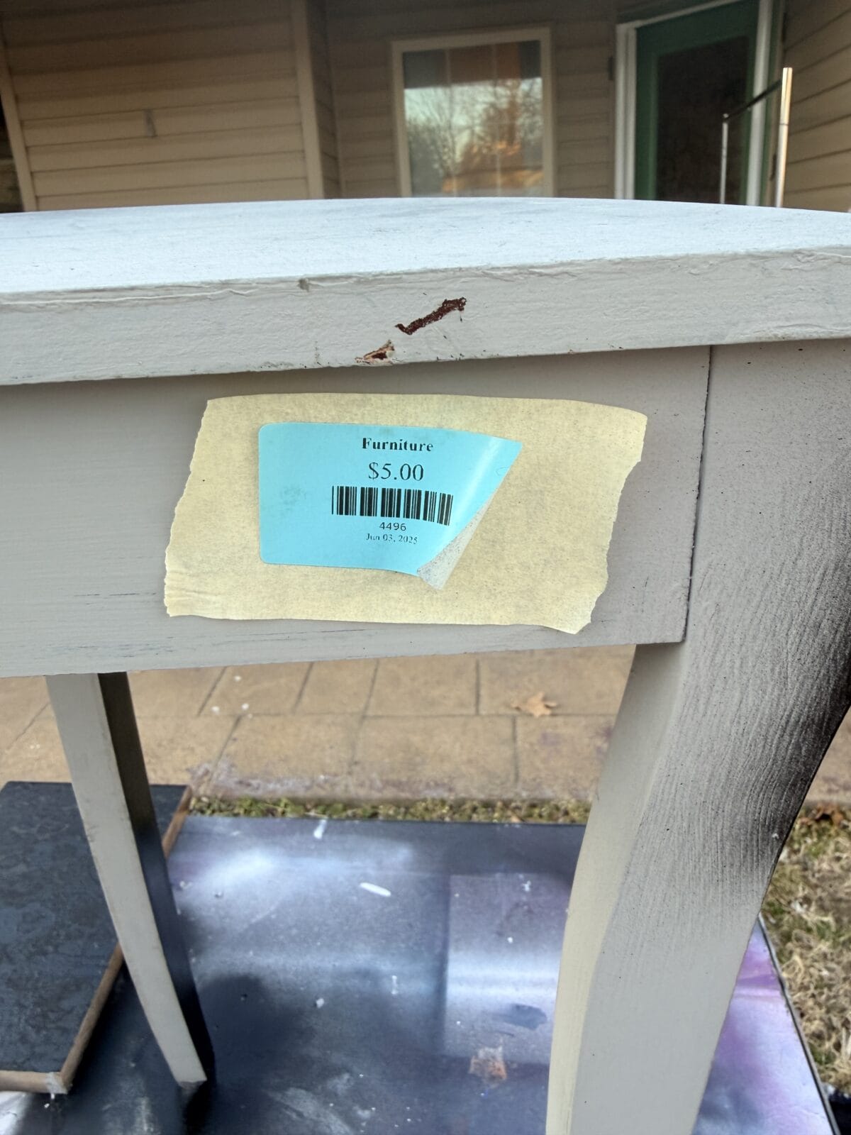A small wooden table with light gray paint and some chipped areas is marked with a blue price tag reading "Furniture $5.00" attached to the side. The table is outdoors on a porch or patio.