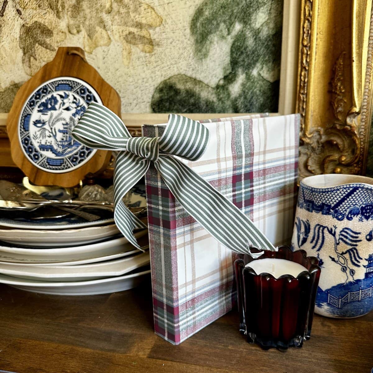 A plaid-wrapped gift with a striped bow sits on a wooden surface beside stacked plates, a blue and white patterned dish, a red glass candle holder, and a decorative mug, with framed art in the background.