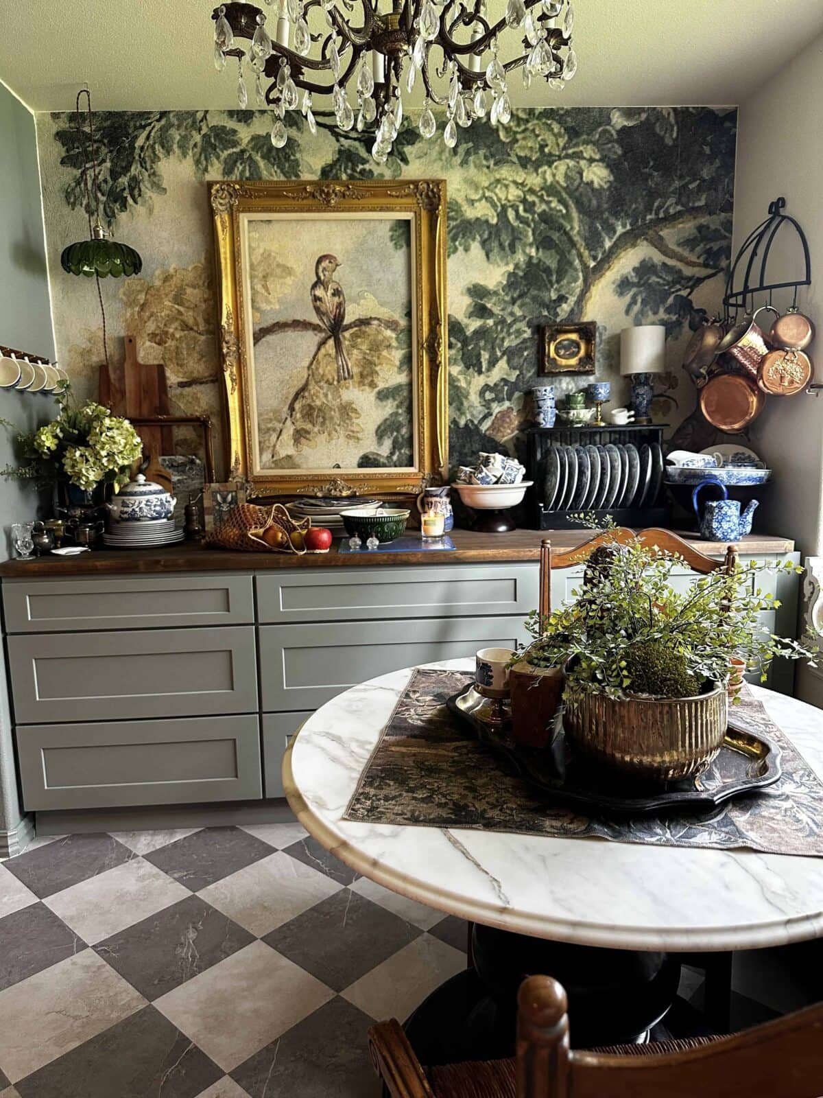 An eclectic kitchen by Amy Sadler Designs features sage green cabinets, a round marble table with potted plants, a framed bird painting, decorative plates, blue and white ceramics, hanging pots, and a chandelier—all set against a nature-themed mural.