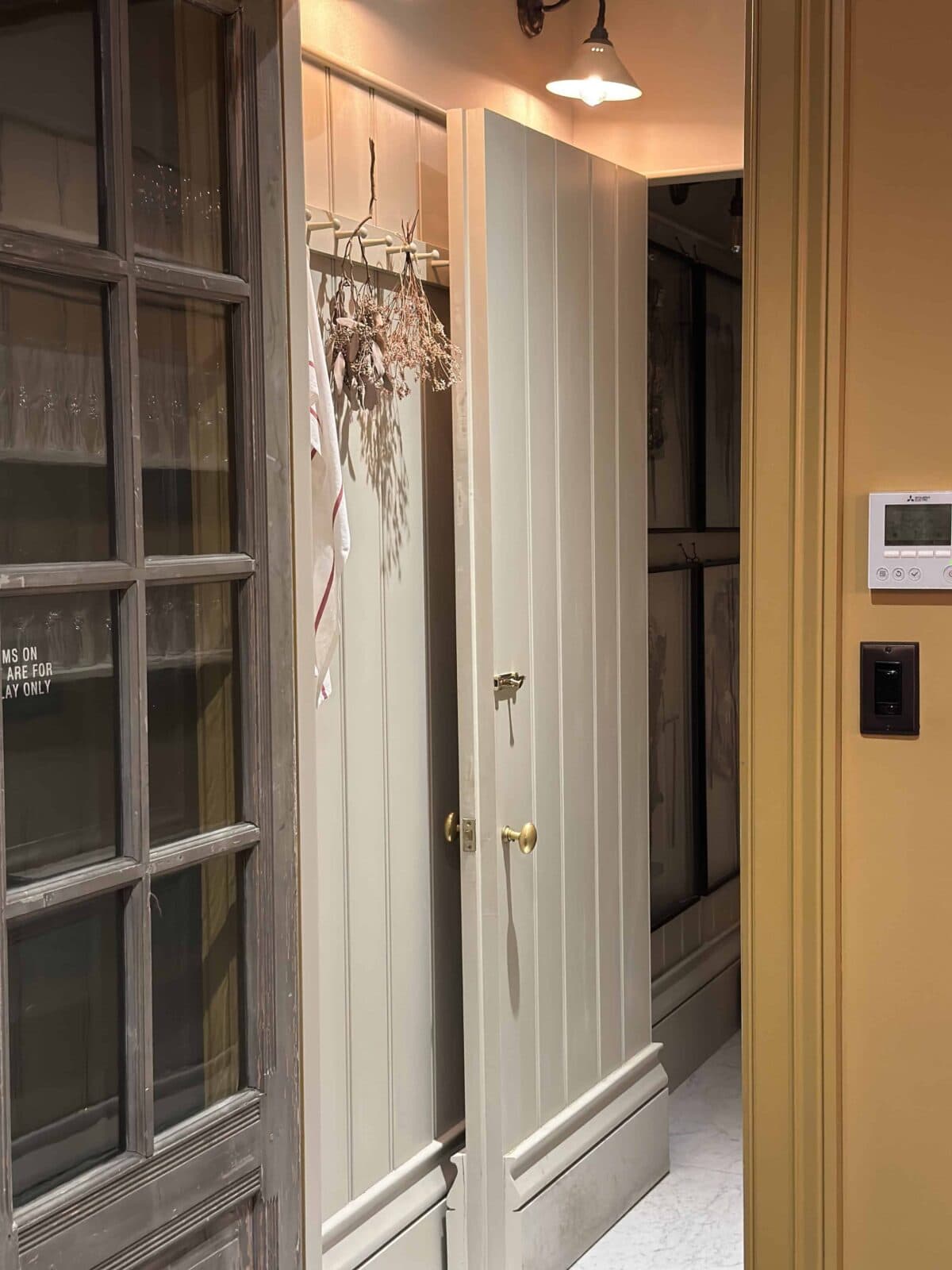 A partially open door reveals a hallway with wall hooks holding dried flowers and striped cloth. There is a glass-panel door on the left and a thermostat with light switches on a mustard yellow wall to the right.