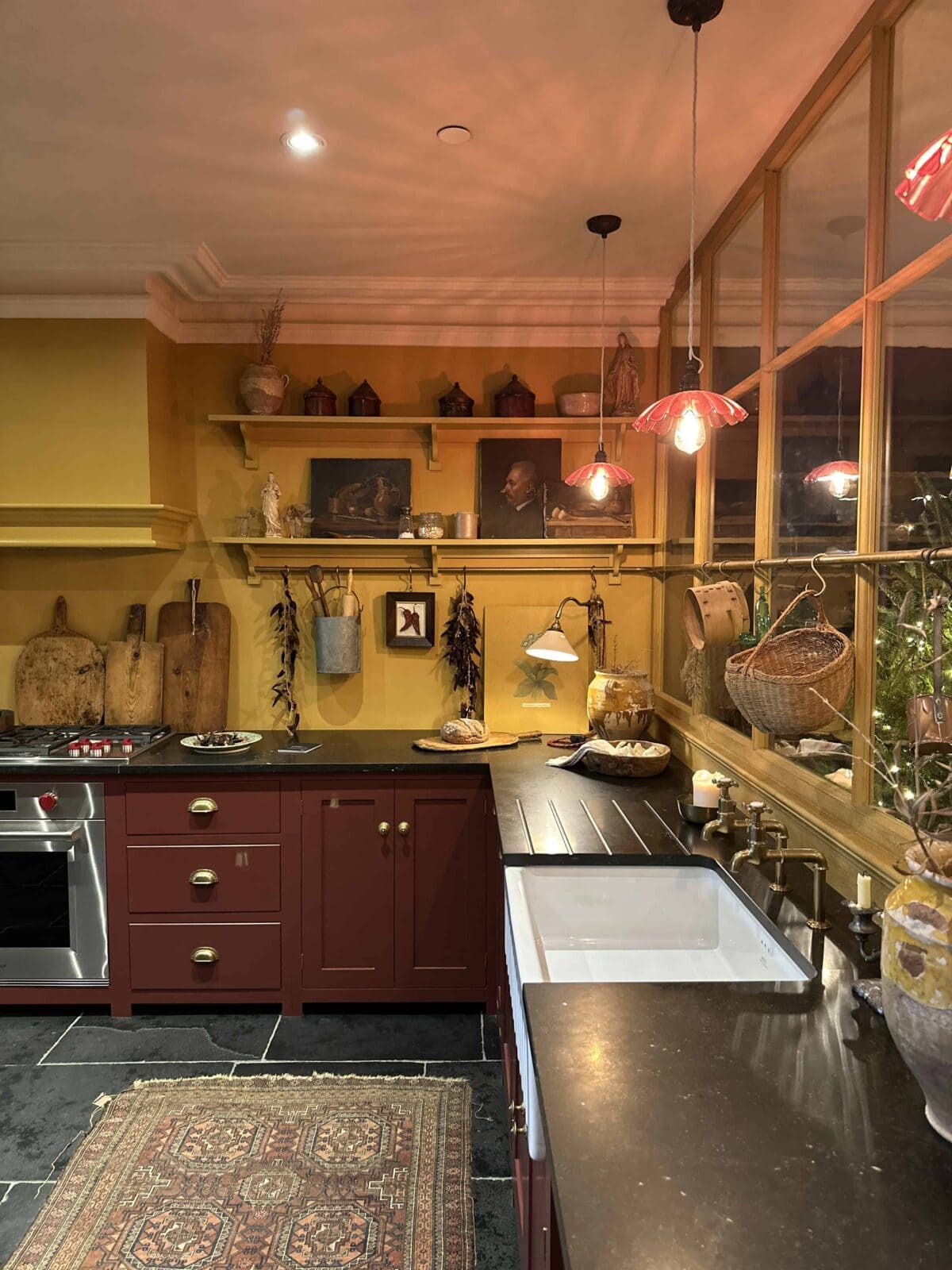 A warm, vintage-style kitchen with mustard yellow walls, burgundy cabinets, a farmhouse sink, open shelves holding decor and kitchen items, hanging red pendant lights, and a patterned rug on a dark stone floor.