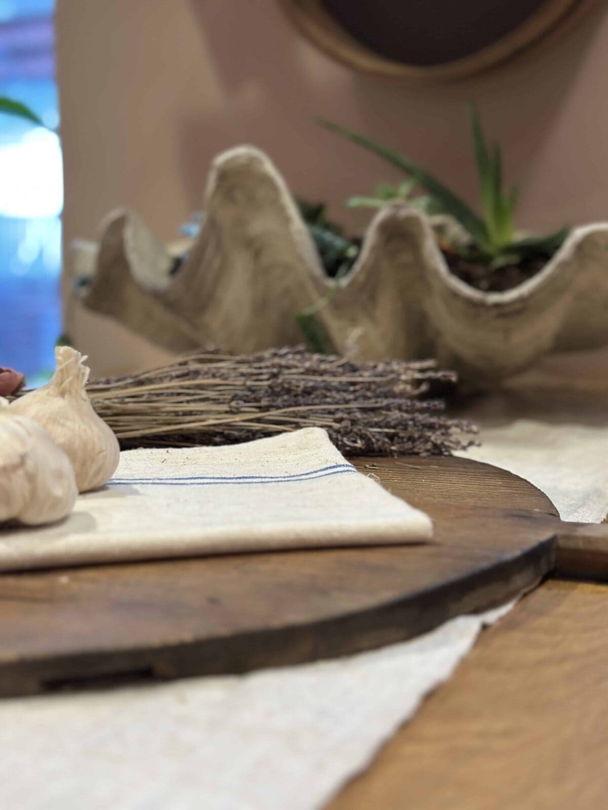 A close-up of a rustic table with a wooden cutting board, a folded white cloth, garlic bulbs, dried lavender, and a decorative clam shell planter with greenery in the background.