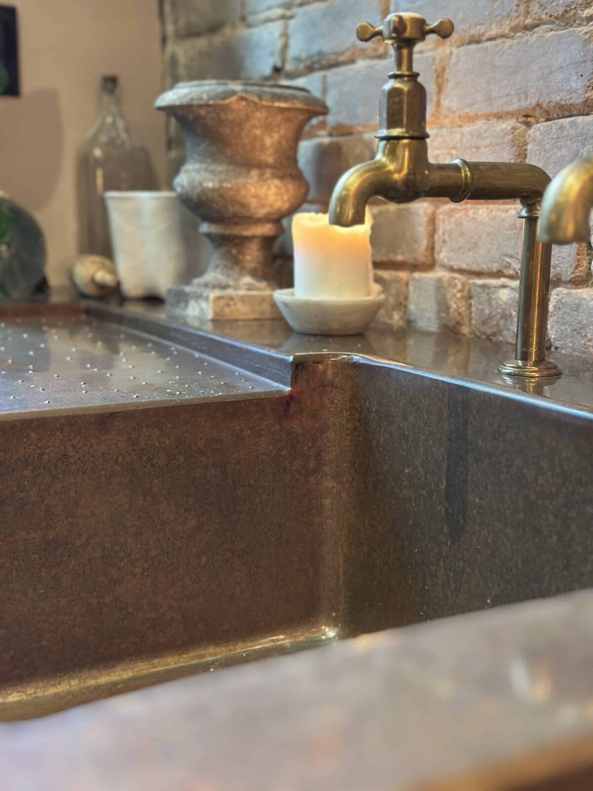 A close-up of a rustic kitchen sink with a bronze faucet, a lit candle, and decorative pottery against an exposed brick wall. The countertop has a metallic texture, and the scene feels cozy and vintage.