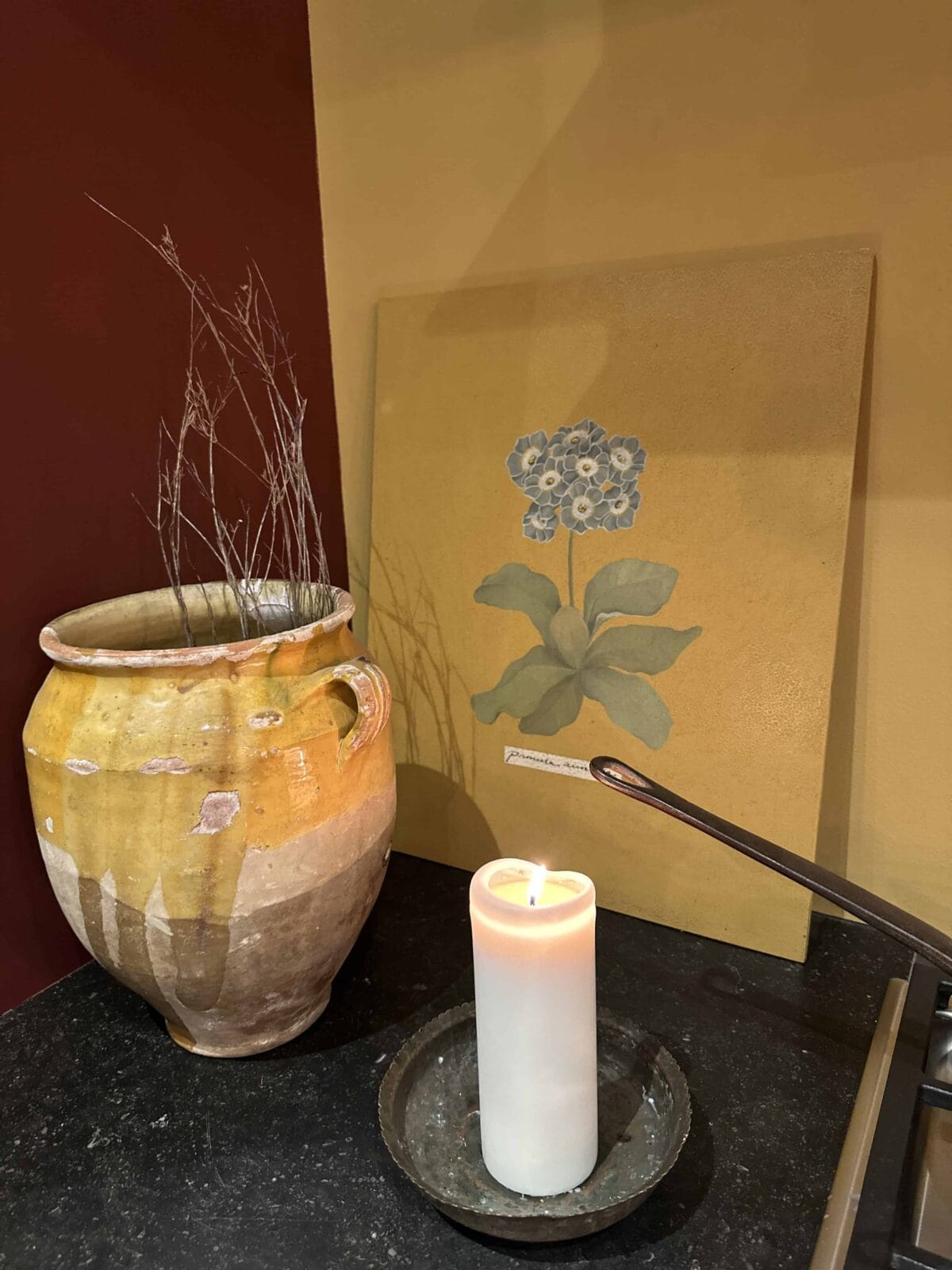 A ceramic vase with dried twigs sits next to a lit white candle and a painting of a blue flower, all arranged on a dark countertop against a yellow and maroon wall.