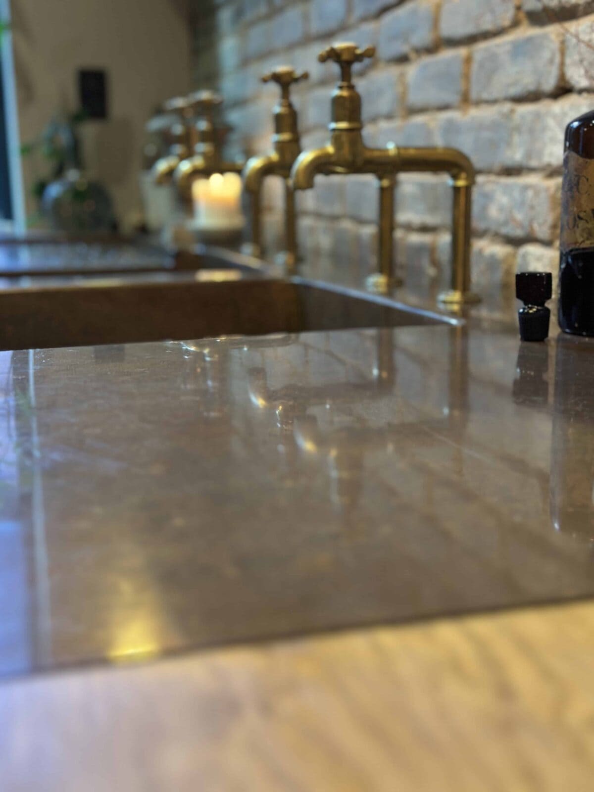 A close-up of a shiny kitchen countertop with brass faucets, a brick wall, a lit candle, and bottles in the background. The focus is on the reflective surface of the counter.