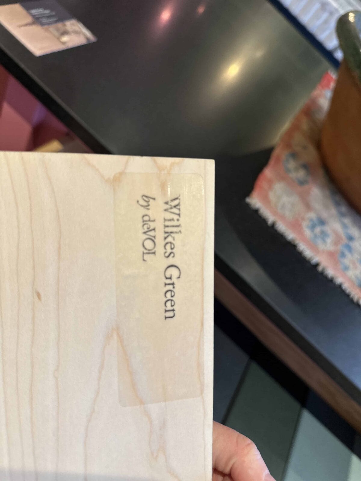 A hand holds a light wooden sample labeled "Wilkes Green by deVOL" over a dark countertop with a colorful patterned rug and a pot partially visible in the background.
