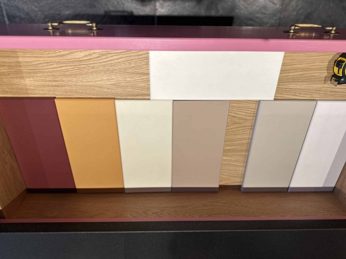 A display of six rectangular color samples in shades of orange, beige, white, and gray, arranged vertically against a wooden background with a pink border. A yellow tape measure is visible on the top right.