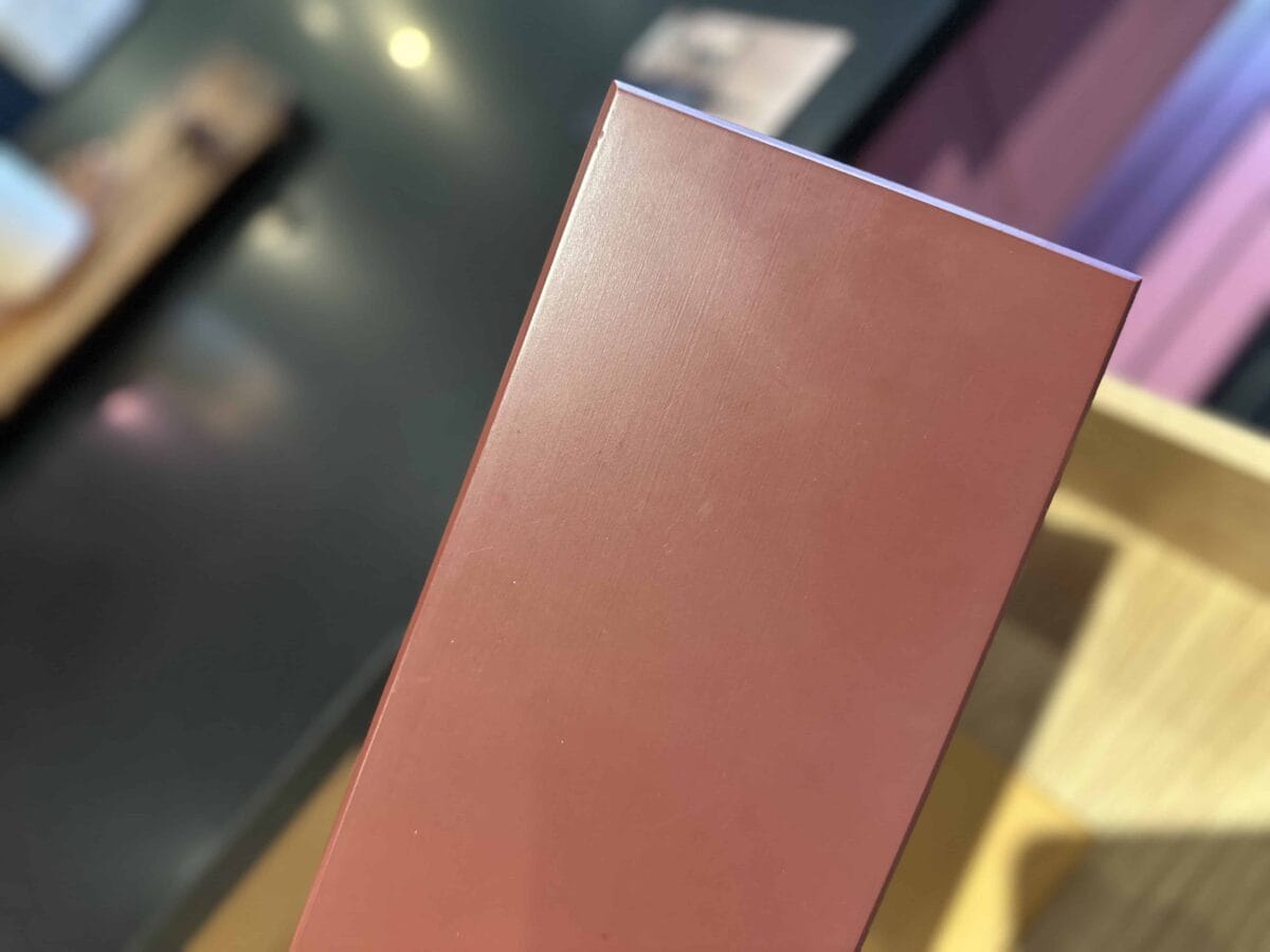 A close-up of a rectangular, dusty pink box held upright, with a blurred background featuring a dark surface and some indistinct objects.
