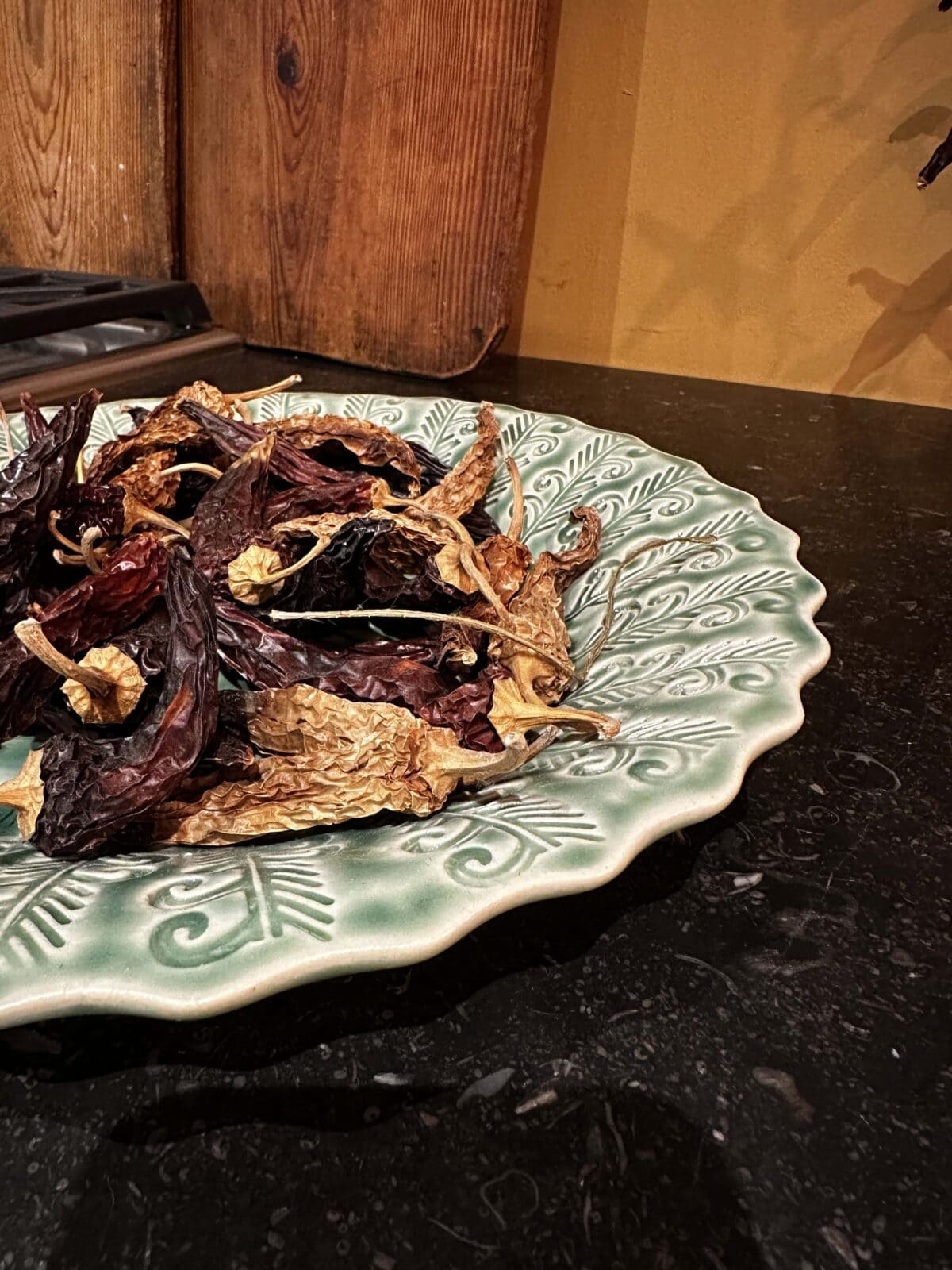 A scalloped green plate with a decorative leaf pattern holds a pile of assorted dried chili peppers on a dark kitchen countertop. Wooden boards and a yellow wall are visible in the background.