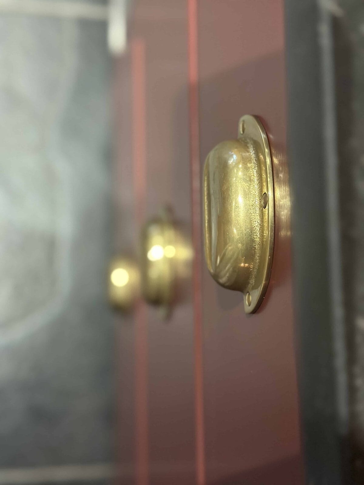 Close-up of shiny brass door handles mounted on a glossy, reddish-brown door, with the background showing another handle out of focus and grey tiled surfaces.