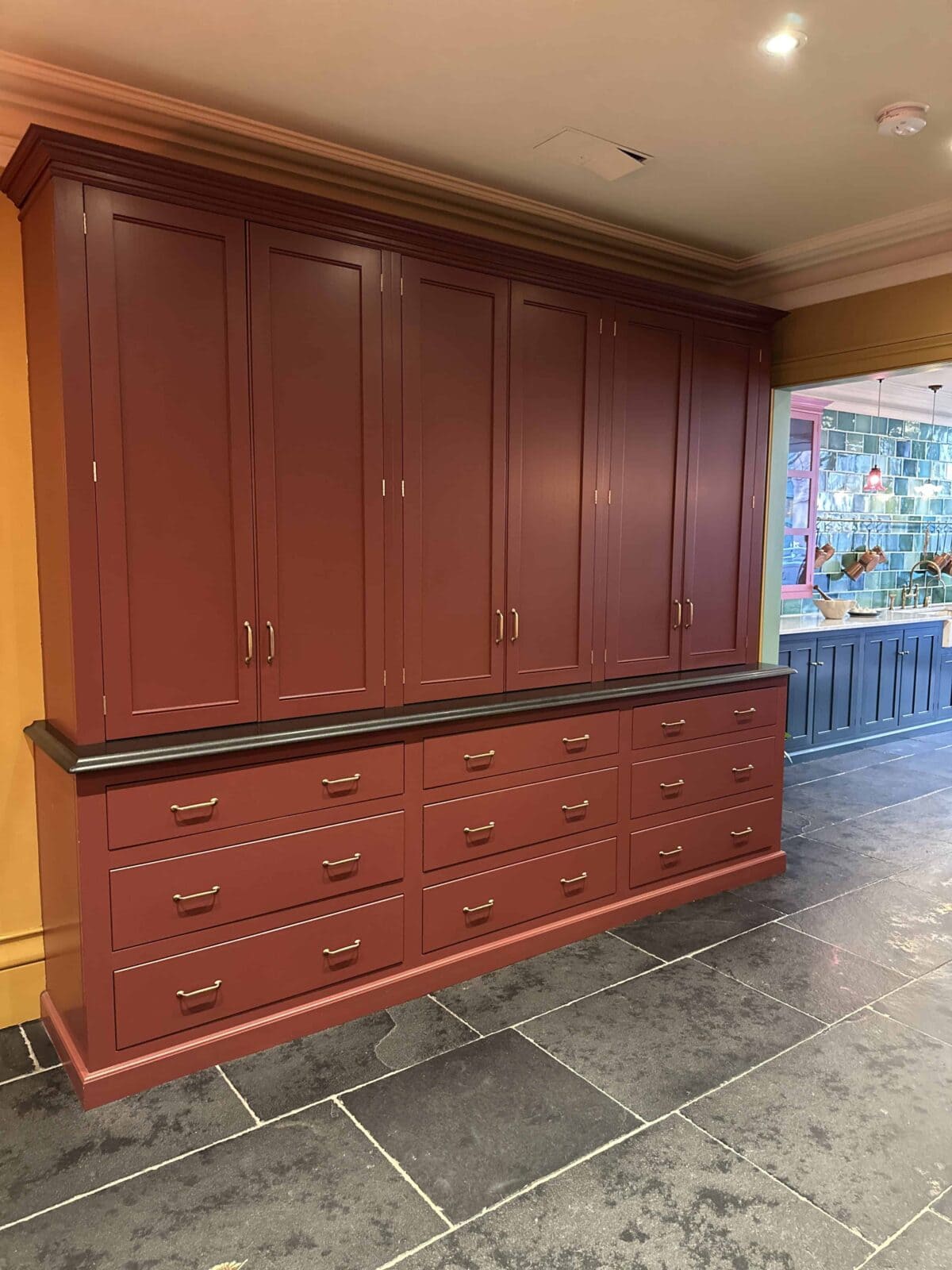 Large built-in cabinet with multiple red drawers and doors, gold handles, and a black countertop, set against a yellow wall with a view into another room featuring more cabinetry and lighting.