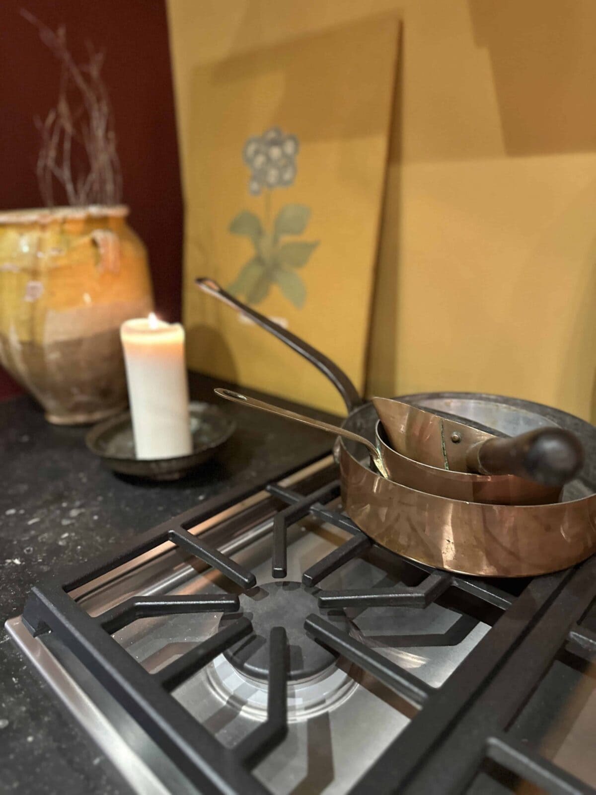 A stovetop with a copper saucepan and utensils resting on it. Beside the stove is a lit white candle on a tray, a yellow ceramic vase, and a yellow floral painting against a dark countertop and reddish wall.
