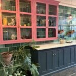 A kitchen with green tiled walls, a blue lower cabinet, pink upper cabinets with glass doors displaying colorful dishes, potted plants, and hanging copper pots. Marble countertop and dark floor tiles are visible.