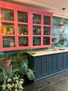 A kitchen with green tiled walls, a blue lower cabinet, pink upper cabinets with glass doors displaying colorful dishes, potted plants, and hanging copper pots. Marble countertop and dark floor tiles are visible.