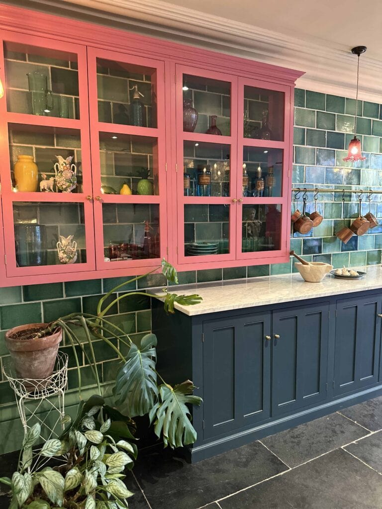 A kitchen with green tiled walls, a blue lower cabinet, pink upper cabinets with glass doors displaying colorful dishes, potted plants, and hanging copper pots. Marble countertop and dark floor tiles are visible.