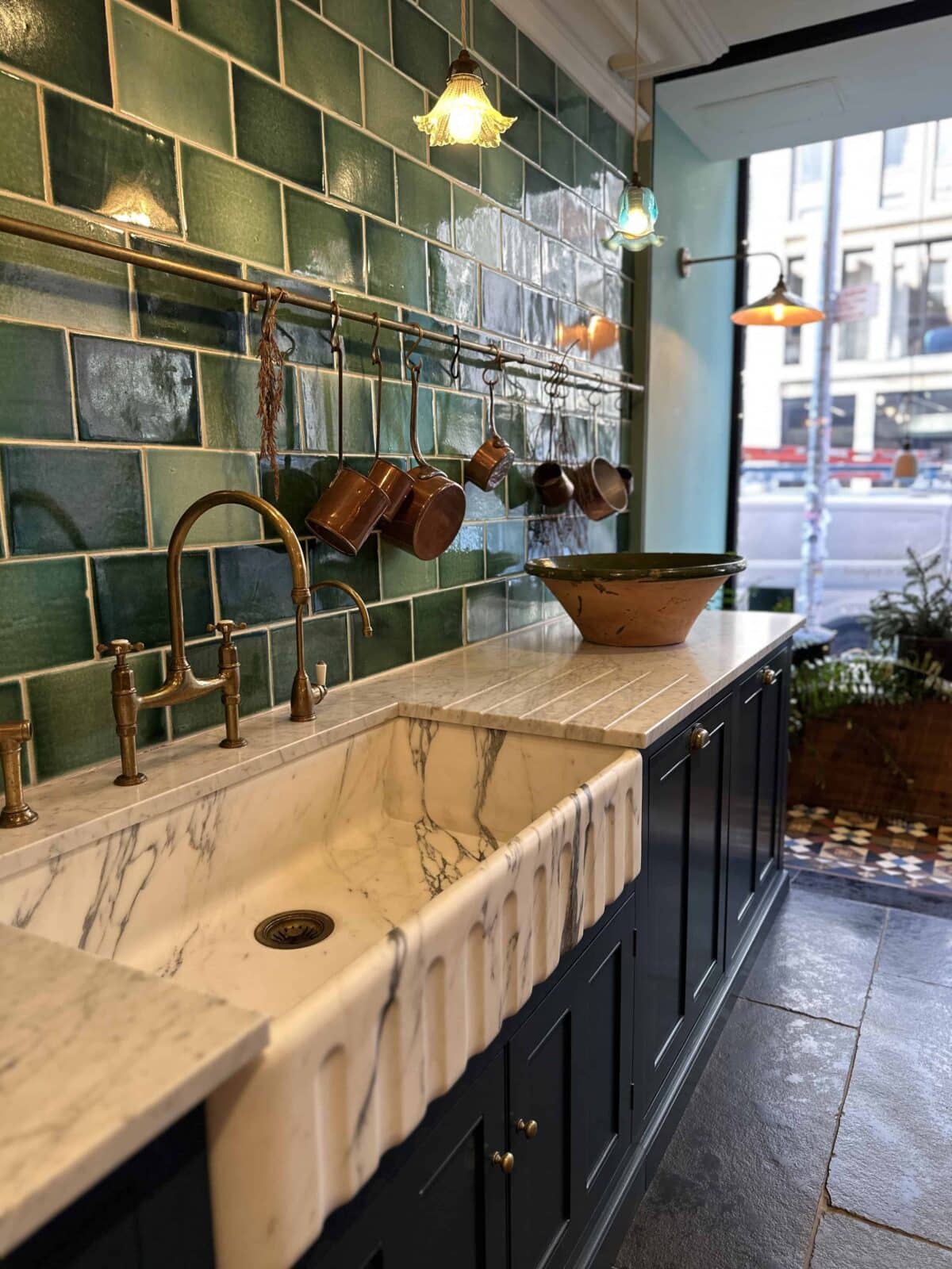 A kitchen with a marble farmhouse sink, brass fixtures, dark cabinets, green tiled backsplash, hanging copper pans, and a large bowl on the counter, next to a window overlooking a city street.