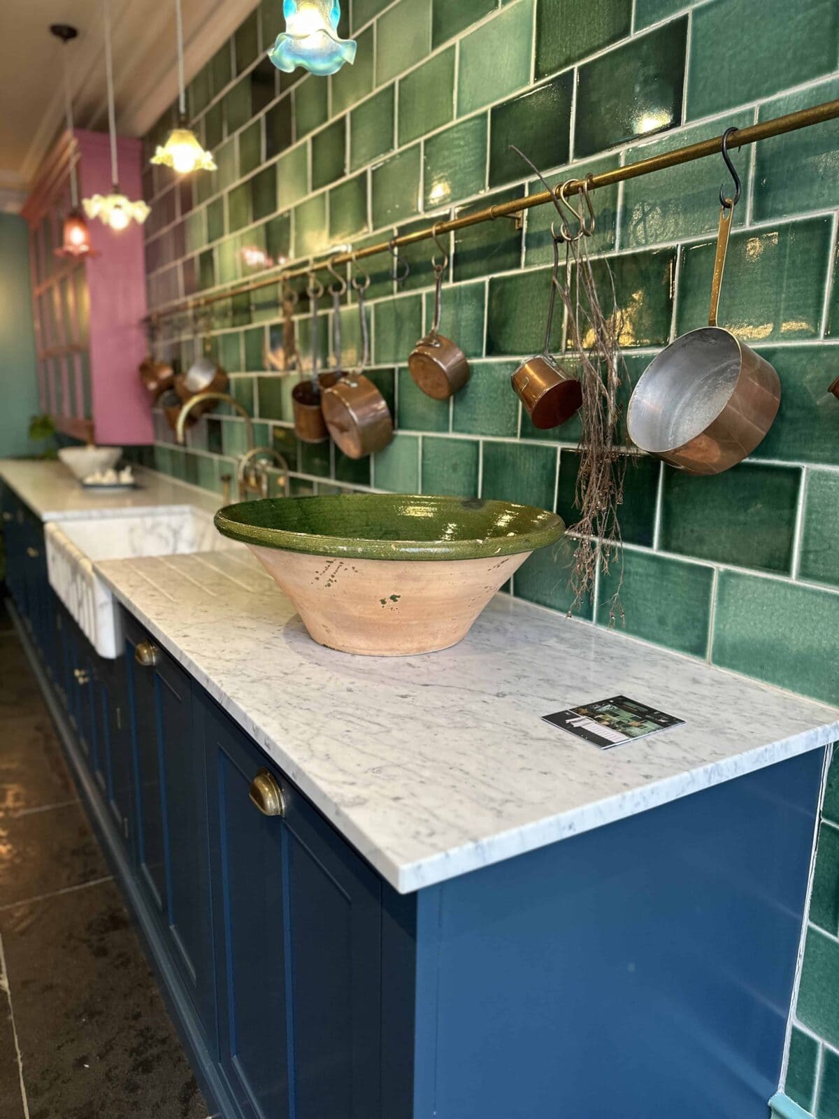 A kitchen with green tiled walls, blue cabinets, a white marble countertop, a large green and tan bowl, copper pots hanging from a rail, and colorful pendant lights.
