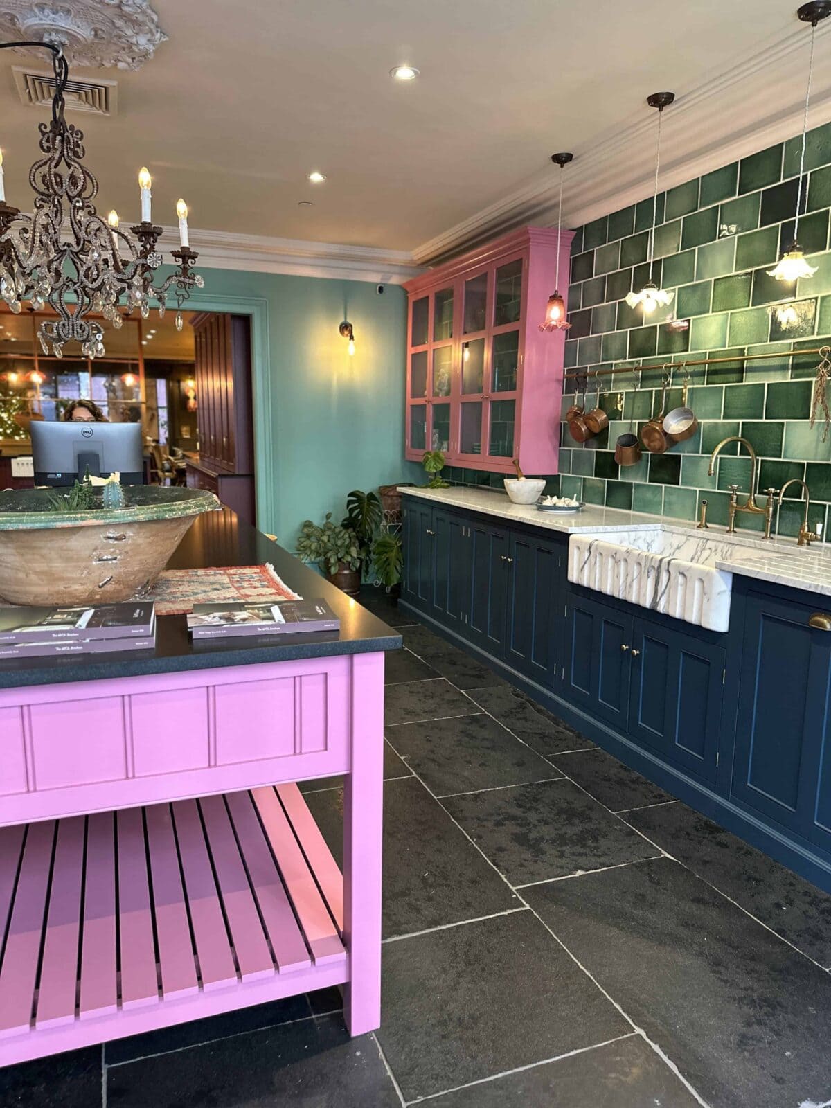 A vibrant kitchen with green tiled backsplash, navy lower cabinets, a pink island, and an ornate chandelier. Copper pots hang above the counter, and the kitchen has a mix of modern and vintage decor.
