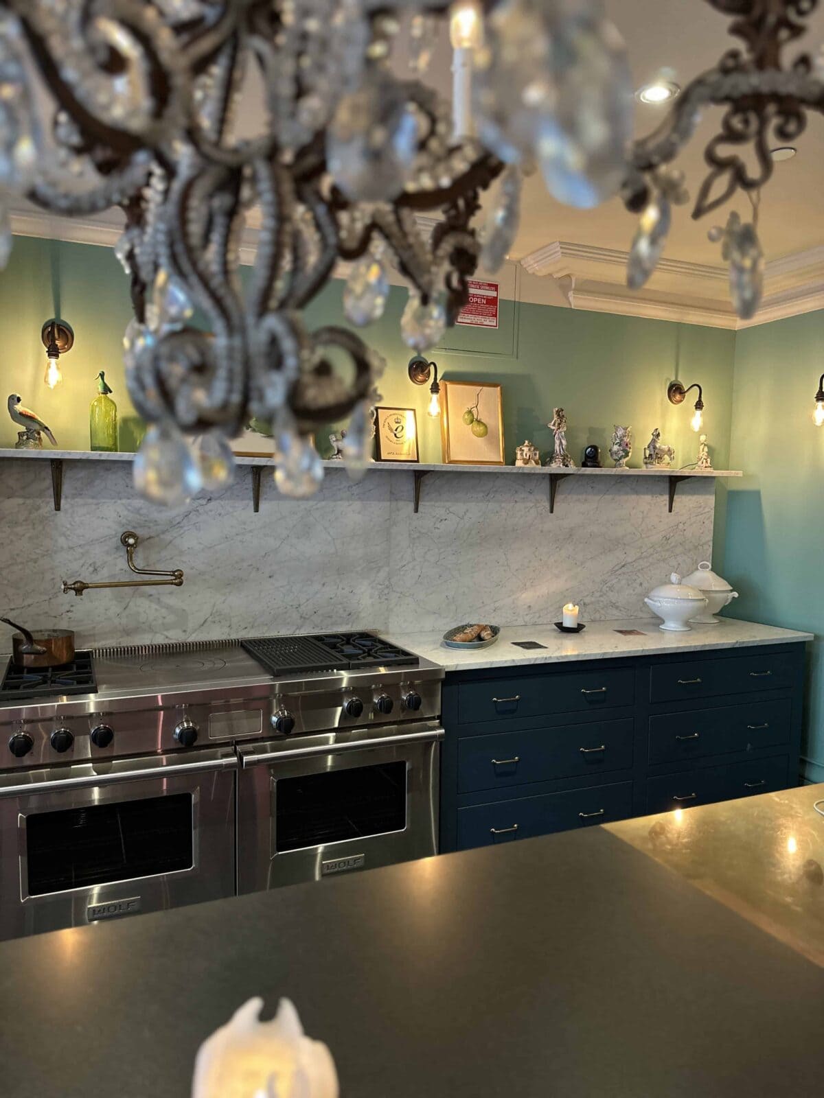 A stylish kitchen with teal cabinets, marble backsplash, a double oven, and decorative items on a shelf. A chandelier with crystal details hangs in the foreground. The room is softly lit with a cozy ambiance.