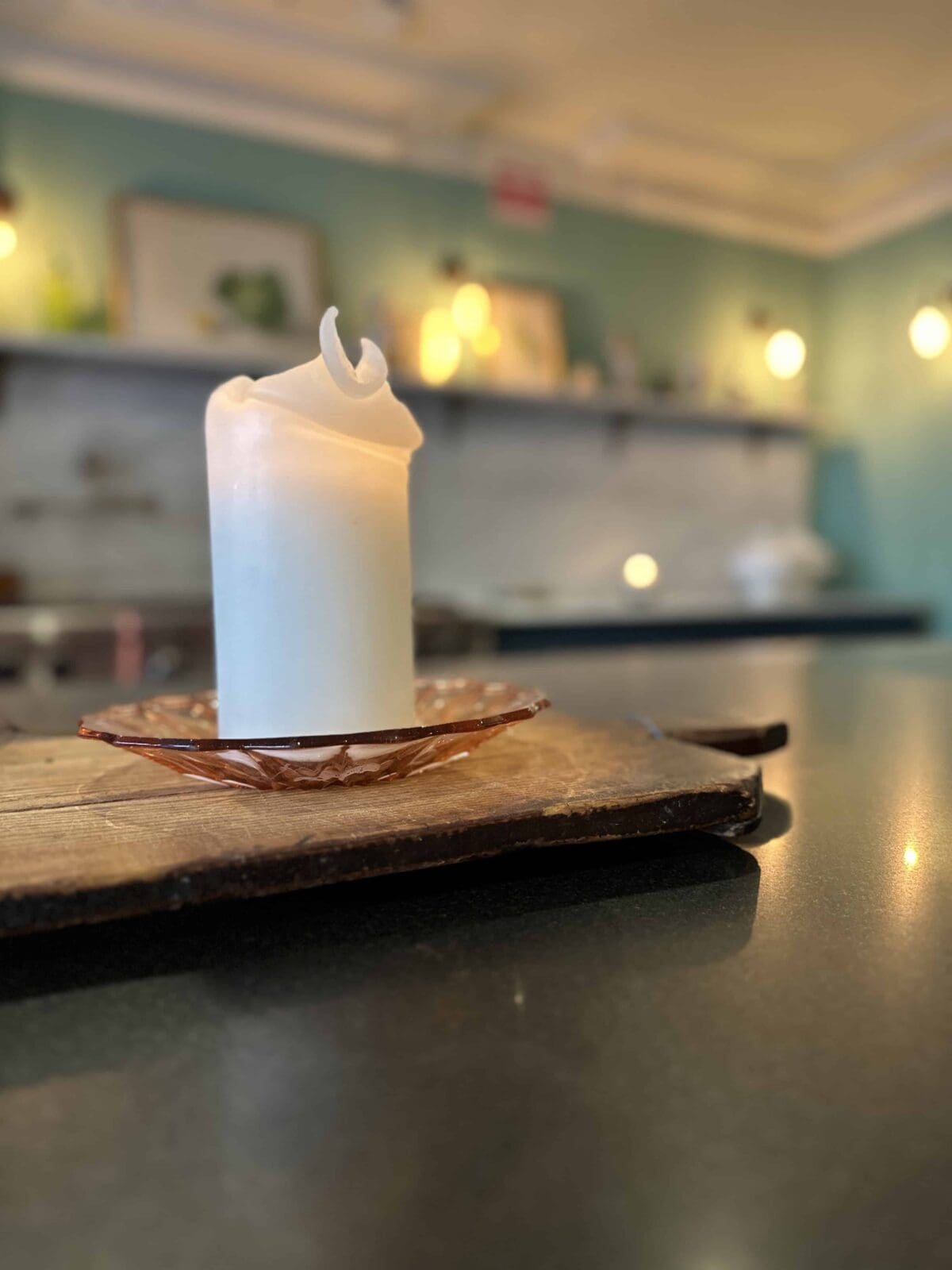 A white candle with a curled wick sits on a glass holder atop a wooden board, with a softly lit, blurred kitchen in the background.