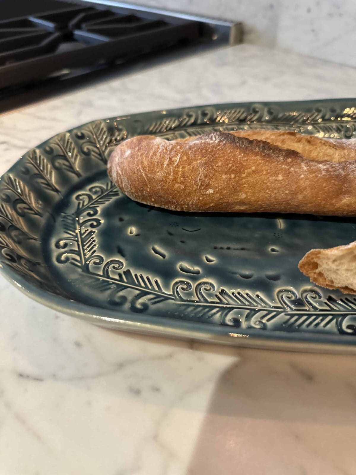 A close-up of a partially eaten baguette on a decorative blue-green plate, placed on a marble countertop near a black stovetop.