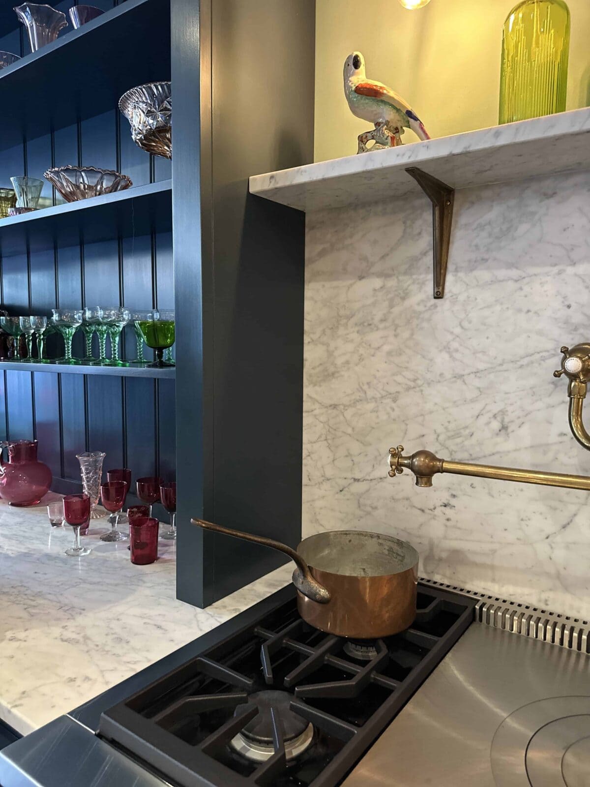 A copper pot sits on a gas stove with water boiling. Behind, there is a marble backsplash, a brass pot filler faucet, and shelves displaying colorful glassware and a ceramic bird.