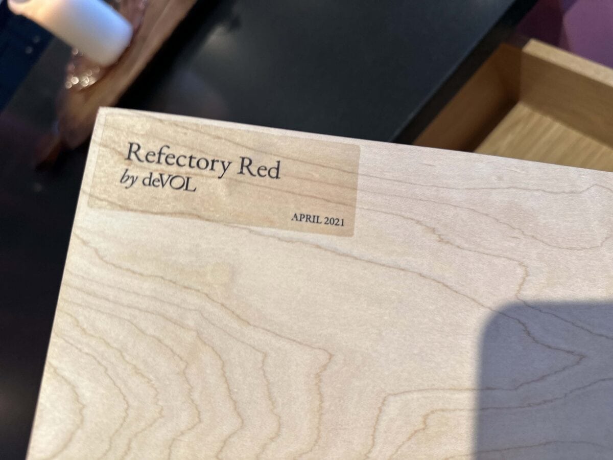 A wooden surface labeled "Refectory Red by deVOL" with "APRIL 2021" printed next to it. Natural wood grain is visible, and part of a candle and wooden tray are seen in the background.