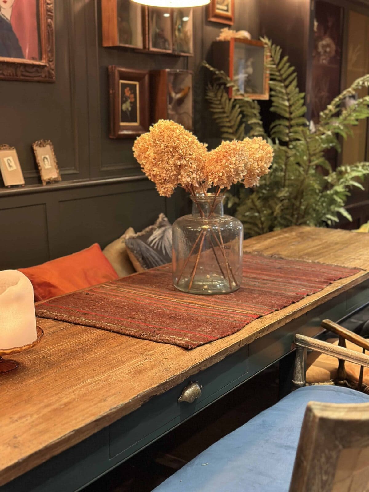 A rustic wooden table with a red runner holds a glass vase filled with dried hydrangeas. The background features framed art, green plants, and warm, cozy decor with orange and blue cushions.