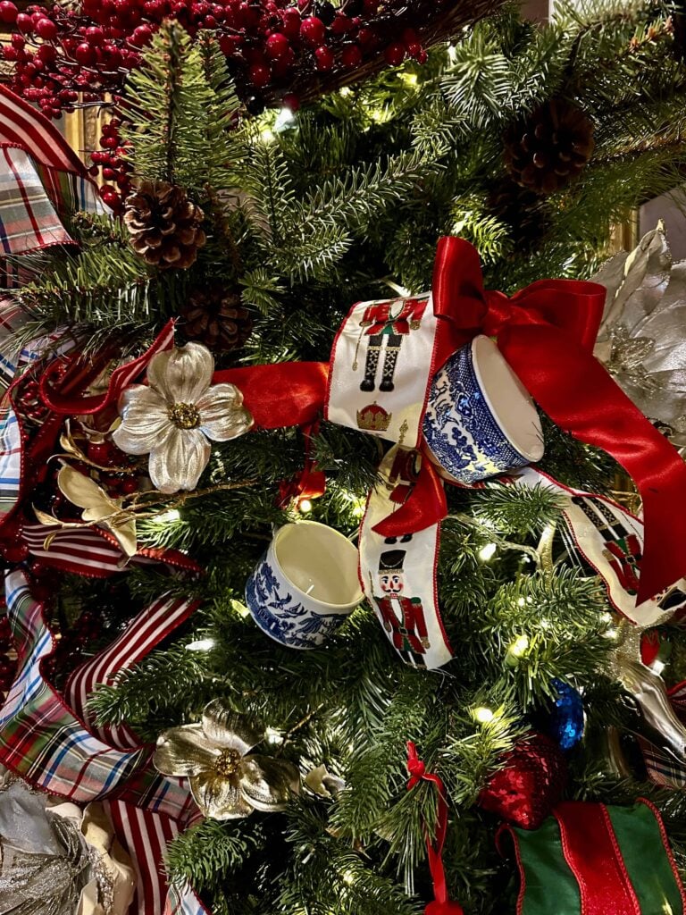 A Christmas tree decorated with red ribbons, gold flowers, pine cones, striped bows, and blue-and-white teacups. The tree is lit with white lights and adorned with festive ornaments and red berries.