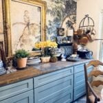 A cozy kitchen with blue cabinets, a wood countertop, and decorative plates is ready for a spring home tour. A large framed bird painting hangs above, with potted plants, yellow flowers, and various kitchenware on the counter and shelves.