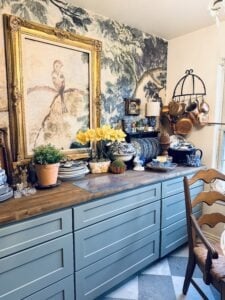 A cozy kitchen with blue cabinets, a wood countertop, and decorative plates is ready for a spring home tour. A large framed bird painting hangs above, with potted plants, yellow flowers, and various kitchenware on the counter and shelves.
