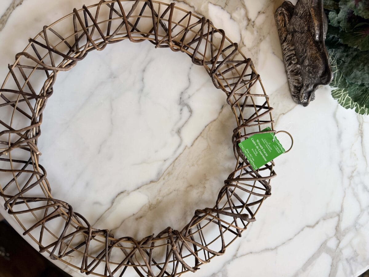 A brown woven wreath with a green tag rests on a white marble surface near a decorative object shaped like an animal's head and some leafy greenery.