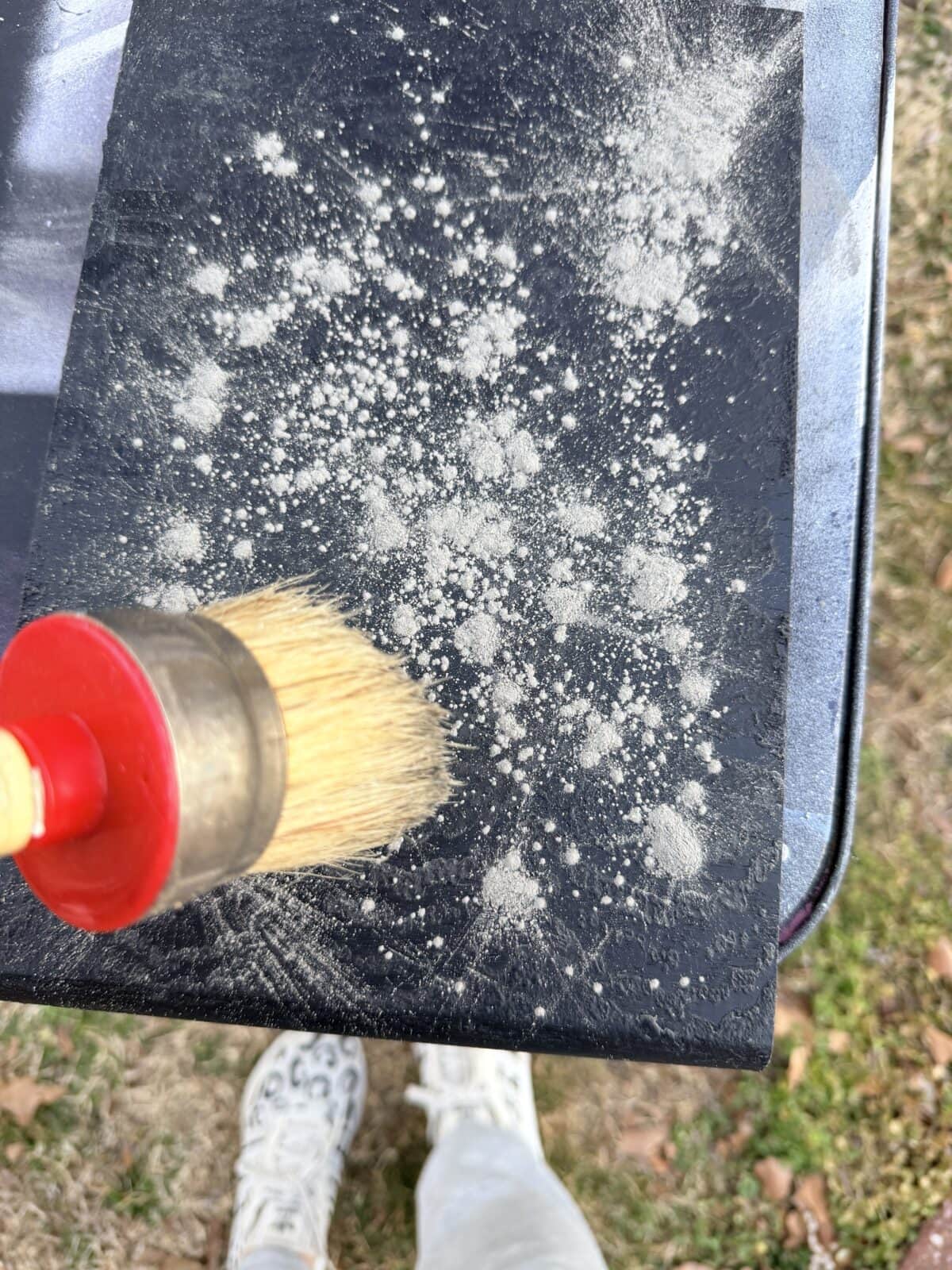 A close-up of a round brush with a red handle spreading white powder on a black surface outdoors. Grass and a person's feet in sneakers and light pants are visible in the background.