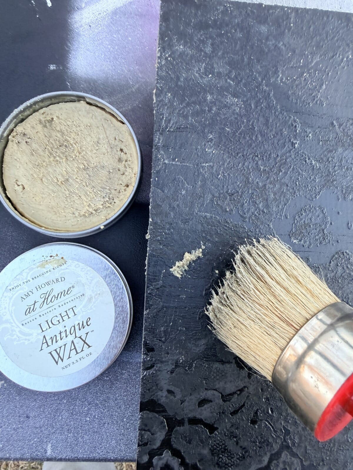 A round tin of Amy Howard at Home Light Antique Wax sits open next to a textured black surface being brushed with wax by a bristle brush with a wooden handle.