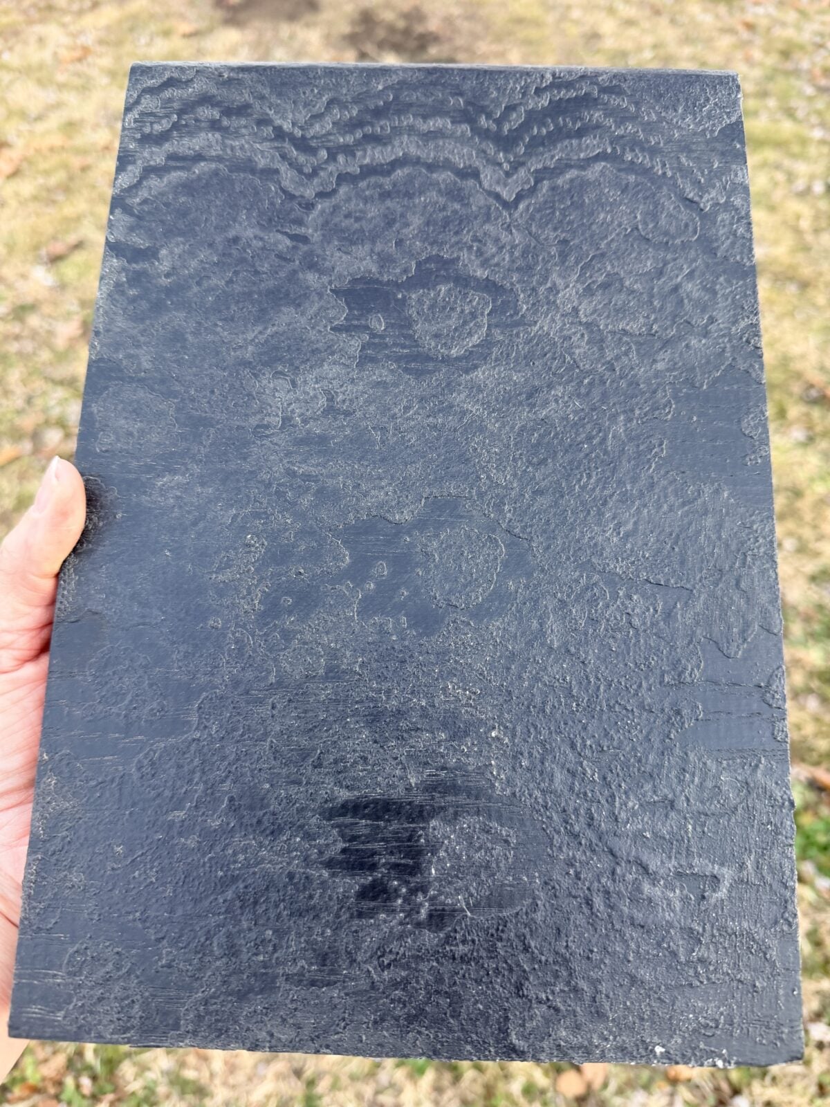 A hand holding a rectangular black stone tile with a textured, wavy surface pattern, outside on grass.