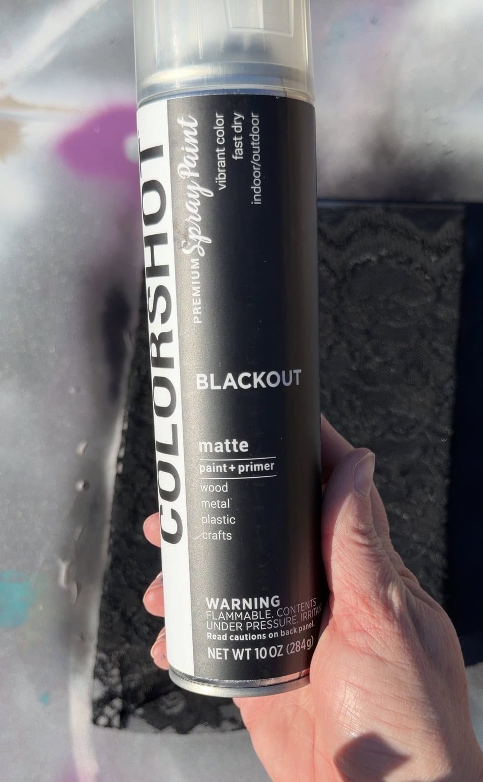 A hand holds a can of COLRSHOT spray paint labeled "BLACKOUT," which is a matte paint and primer for wood, metal, plastic, and crafts. Sunlight casts shadows across the label.