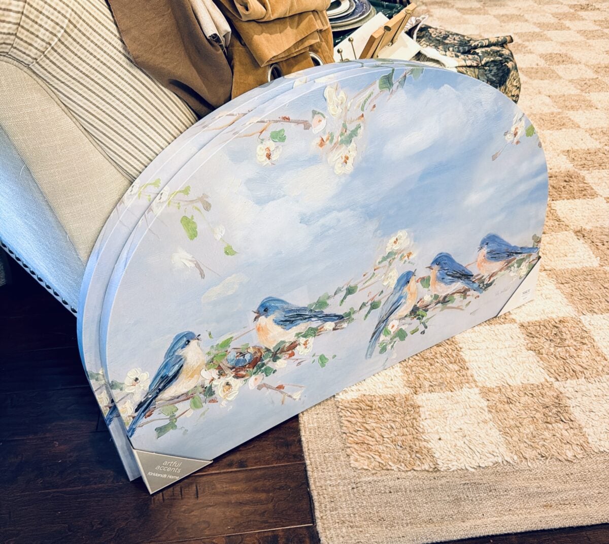 A semi-circular board with a painted design of bluebirds sitting on flowering branches rests against a beige couch on a checkered rug, adding charm to your spring home tour amidst various nearby items.