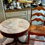 A small round table with a vintage world map design on top sits next to a wooden chair with a woven seat. Behind, a counter holds decorative items, flowers, and china, with framed art on the wall above.