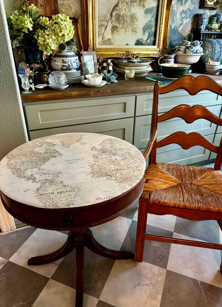 A small round table with a vintage world map design on top sits next to a wooden chair with a woven seat. Behind, a counter holds decorative items, flowers, and china, with framed art on the wall above.