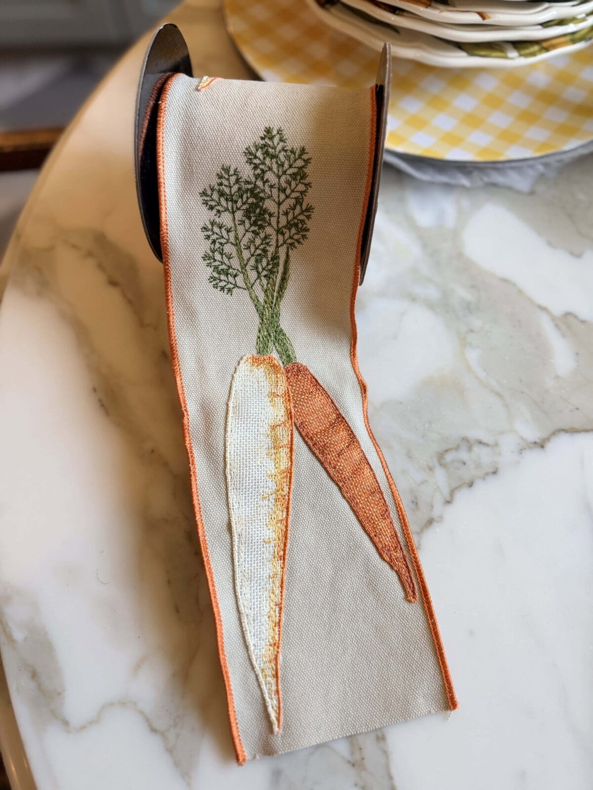 A spool of beige ribbon with orange edges, featuring embroidered images of two carrots with green tops, sits on a marble tabletop near a stack of yellow checkered plates.