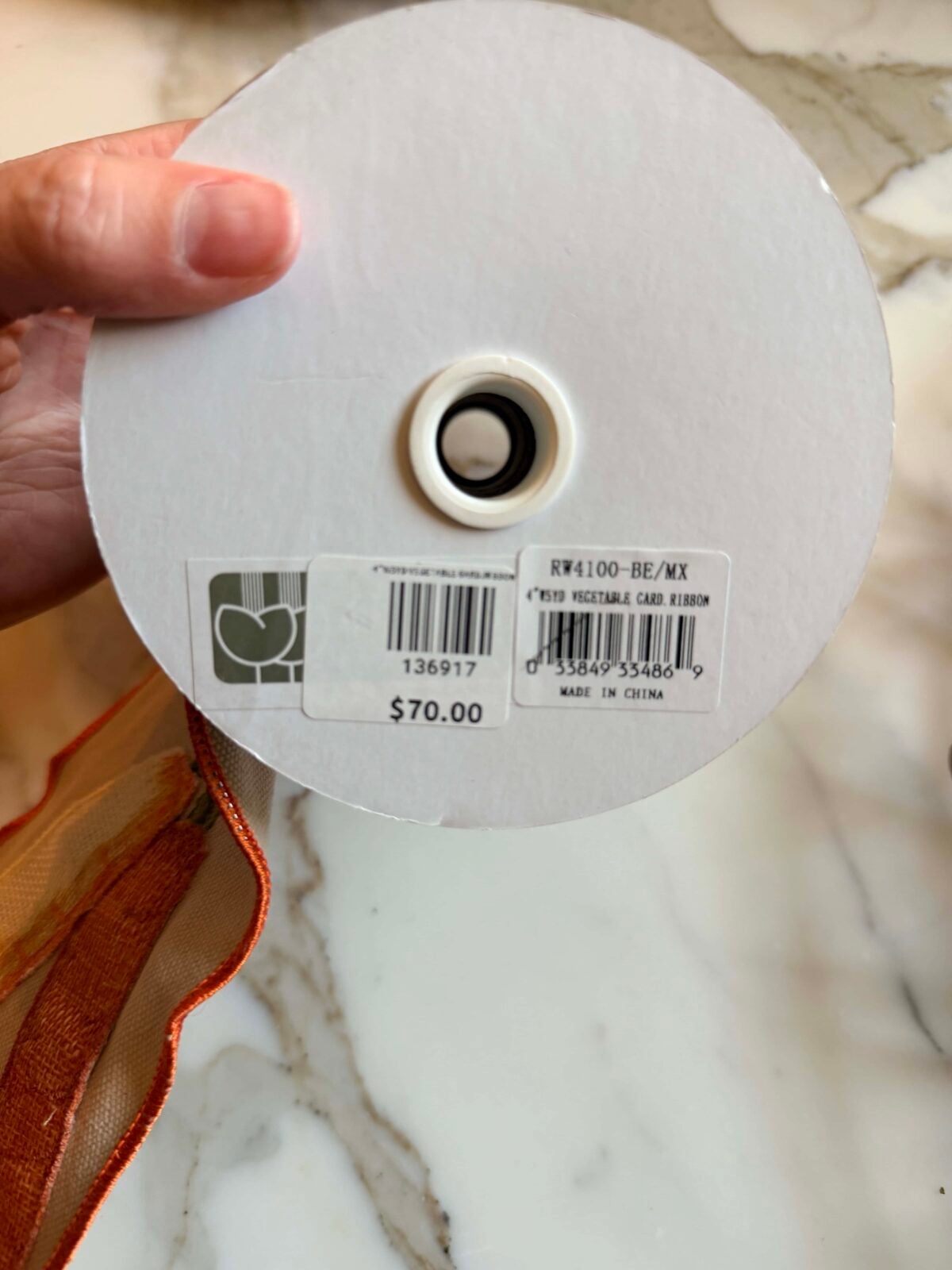 A hand holds a spool of ribbon with a white label showing a barcode, numbers, and a price of $70. The ribbon is orange with a decorative edge, and the background is a marble surface.