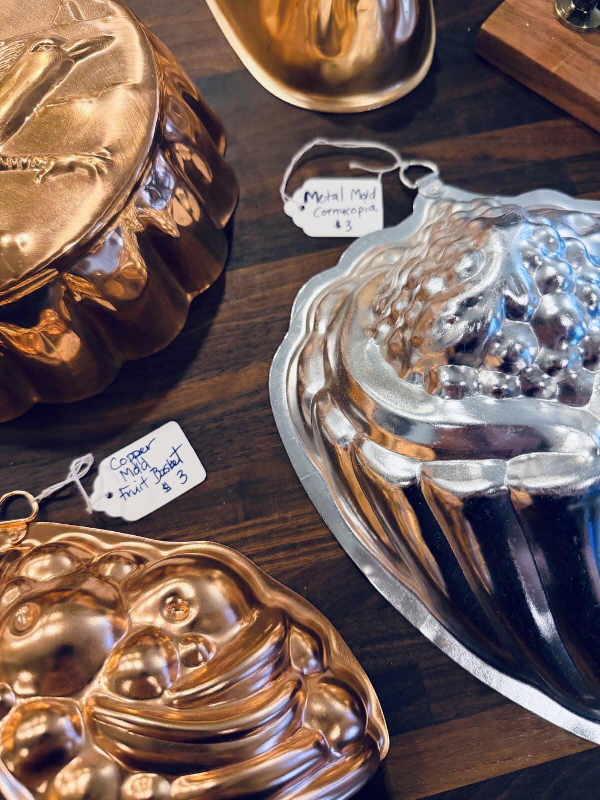 A close-up of various vintage copper and metal baking molds displayed on a wooden surface, each with handwritten price tags—perfect accents for your spring home tour, featuring detailed fruit and cornucopia designs.