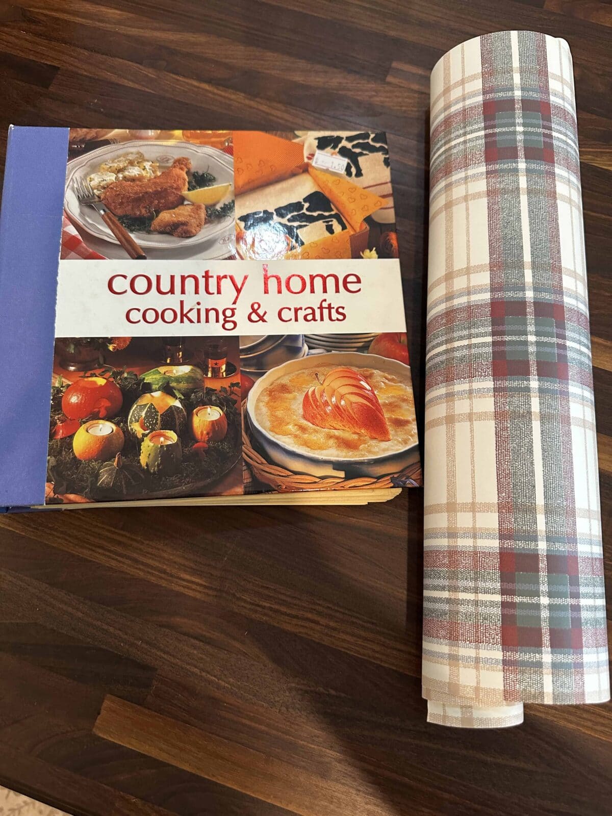 A cookbook titled "country home cooking & crafts" sits on a dark wooden surface next to a roll of red, green, and white plaid-patterned paper.
