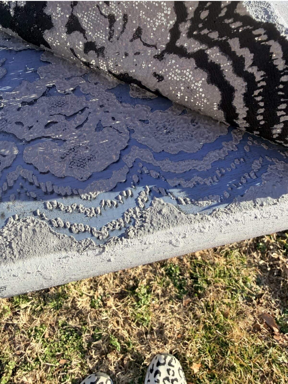 A close-up of layered patterned fabrics in shades of gray, black, and white, with floral and abstract designs, resting on a grassy surface. Part of a person’s leopard-print shoes are visible at the bottom.