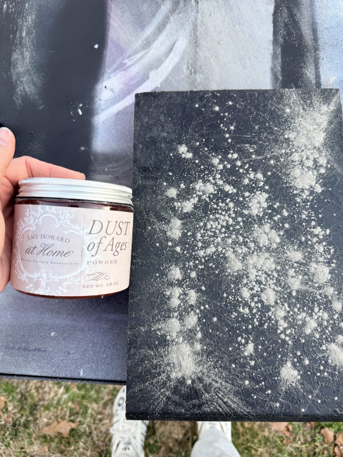 A hand holds a tin labeled "Dust of Ages Powder" next to a black rectangular board sprinkled with powdery dust. Grass and shoes are visible in the background.