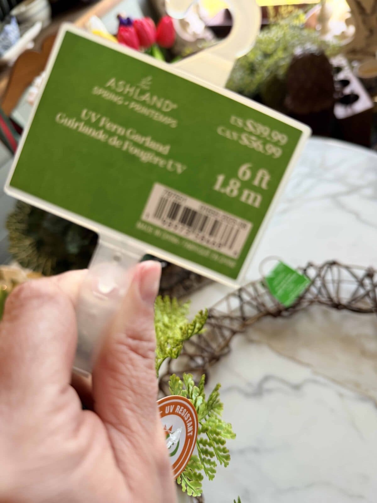 A hand holds a price tag for an Ashland UV Fern Garland, 6 feet (1.8 meters), priced at $39.99 USD or $52.99 CAD, near artificial greenery on a marble surface.