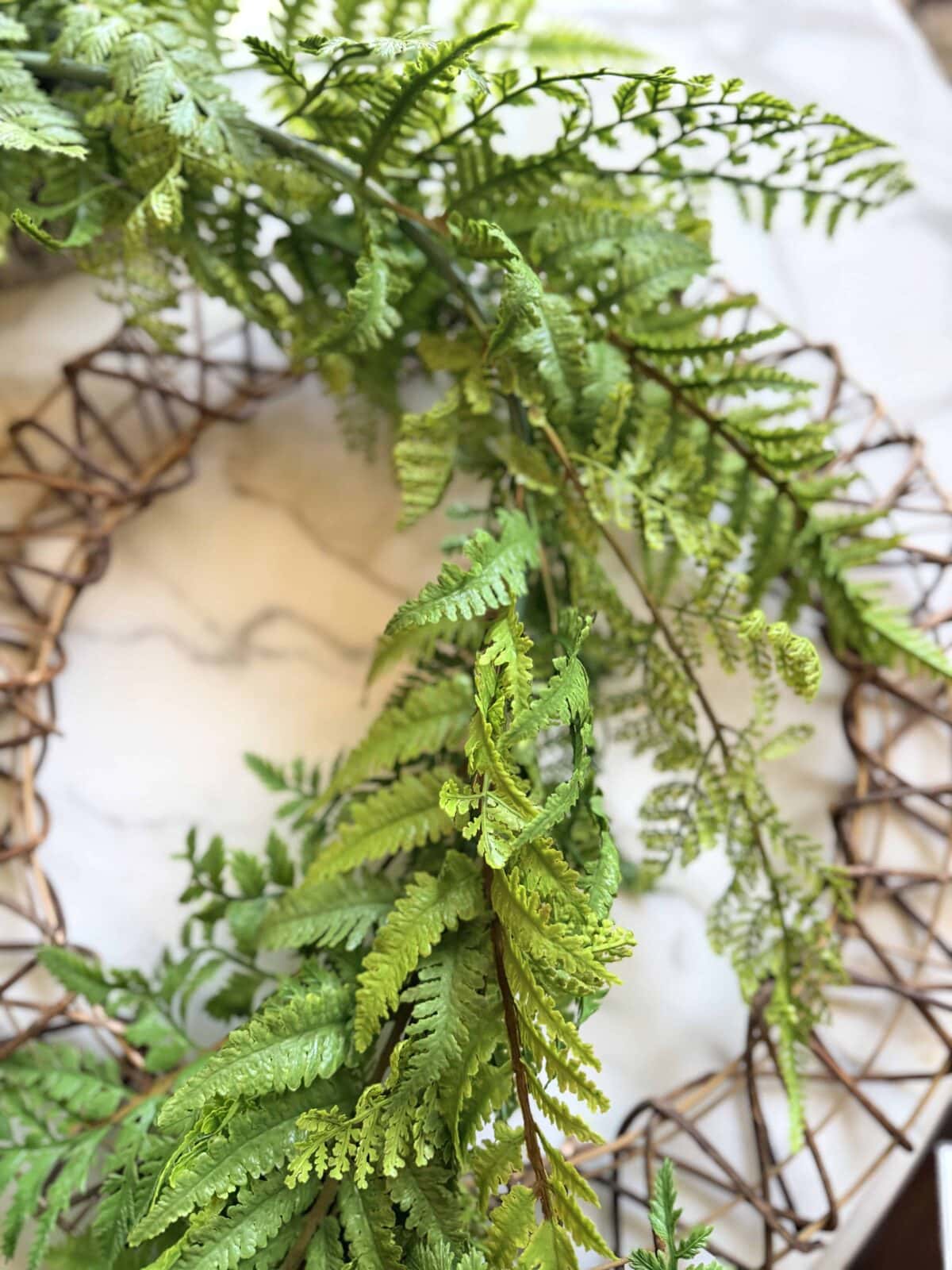 A close-up of a wreath made from woven brown twigs, decorated with vibrant green fern leaves, resting on a white marble surface.