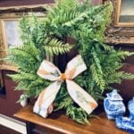 A green fern wreath with a white and orange ribbon bow sits on a wooden table, surrounded by framed paintings on a maroon wall and blue-and-white ceramics—perfect for your spring home tour.