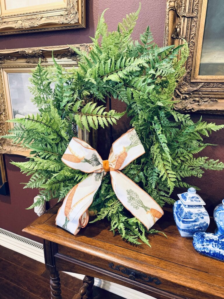 A green fern wreath with a white and orange ribbon bow sits on a wooden table, surrounded by framed paintings on a maroon wall and blue-and-white ceramics—perfect for your spring home tour.